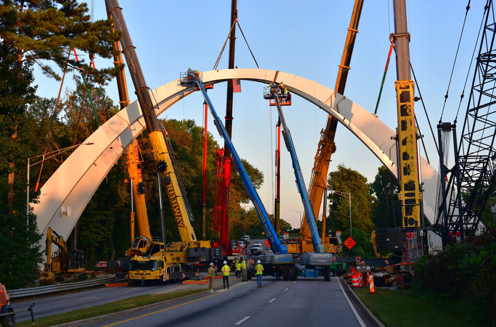 Construction crews and five large cranes work in tandem to lift and assemble segments of a white steel arch over a closed roadway during the erection of the Global Gateway Connector Bridge in College Park, Georgia.