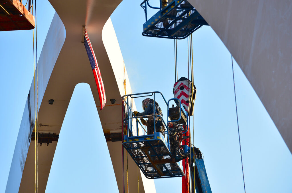 A construction worker in full protective gear welds a steel joint on the white arch of the Global Gateway Connector Bridge.