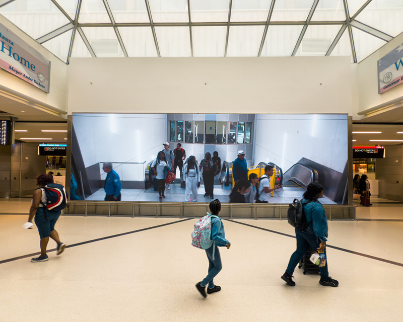 People walk past the large screen showing the arrival of passengers to the terminal from the plane train.