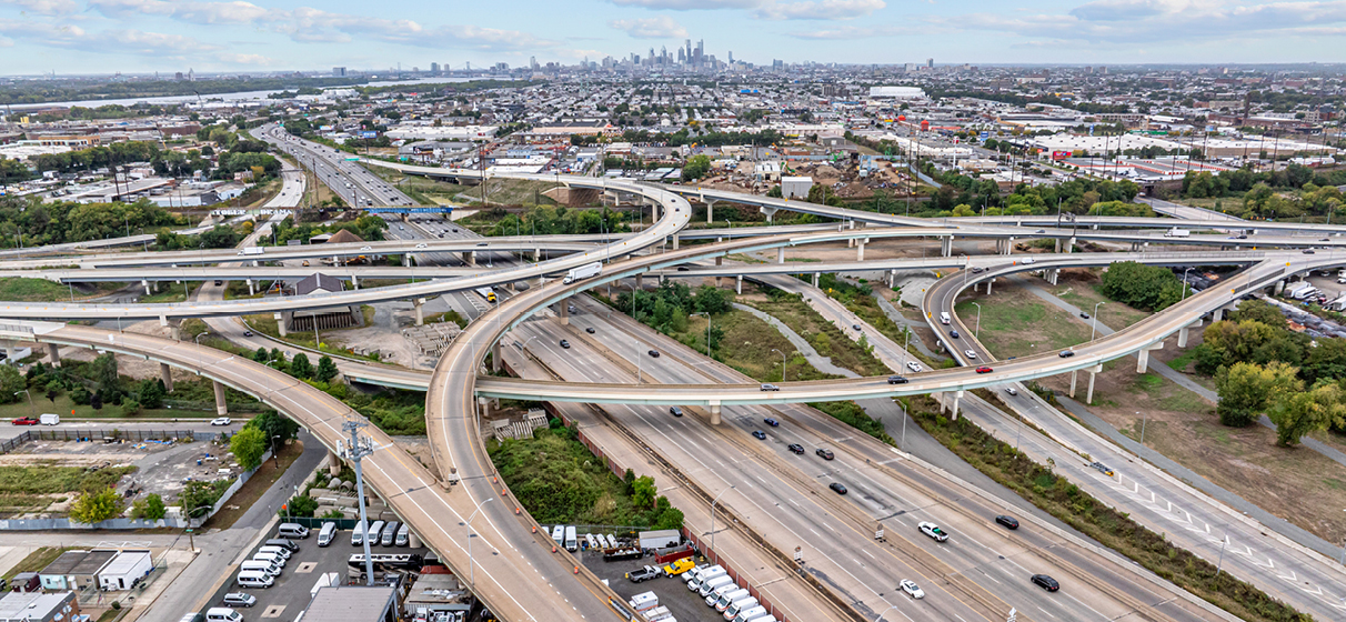 Aerial photo of the Betsy Ross interchange with bridges and ramps crossing over a highway with houses, businesses, and the Philadelphia skyline in the background.