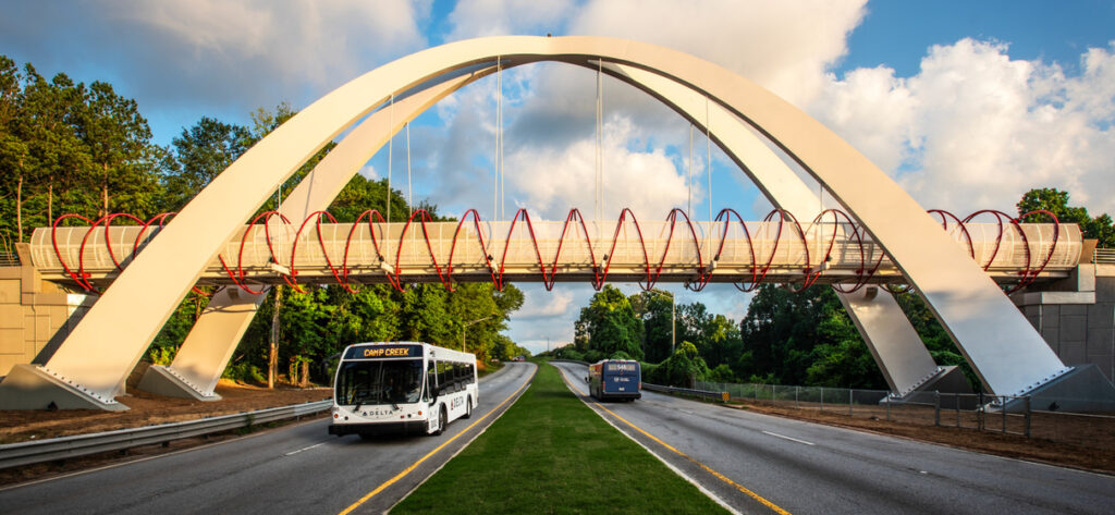 Ground-level view of Global Gateway Connector Bridge crossing a roadway with a bus and car passing underneath and an airplane flying above it.
