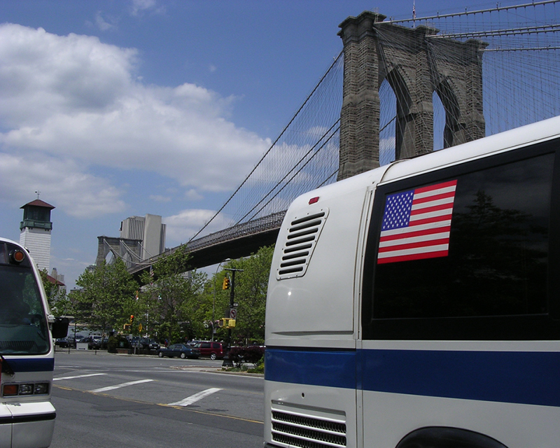 NYC busses await passengers with the Brooklyn Bridge in the background.