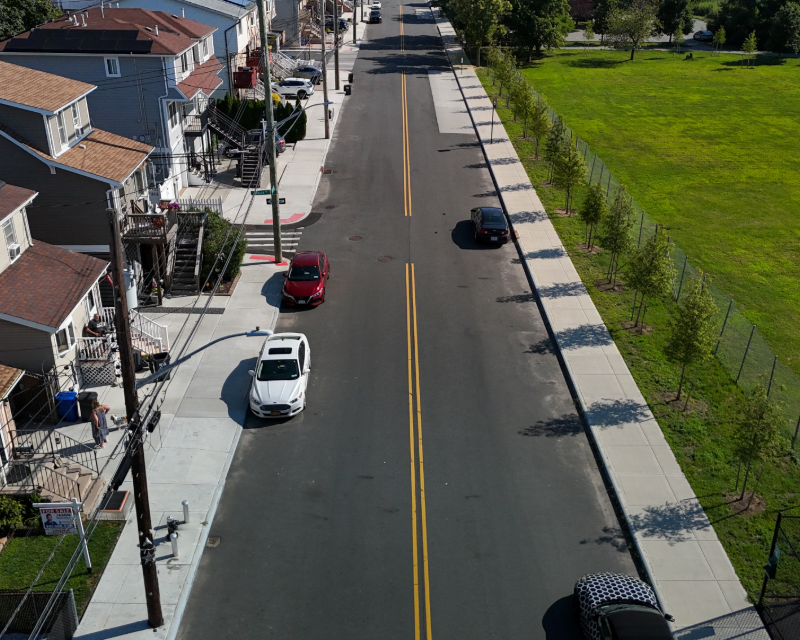 Aerial of Staten Island roadway with cars parked on street, one side lined with houses, the other trees and grass.
