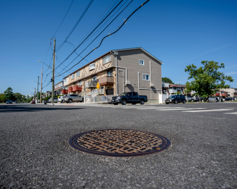 Close up of sewer manhole cover on street with home in the background in Staten Island, NY.