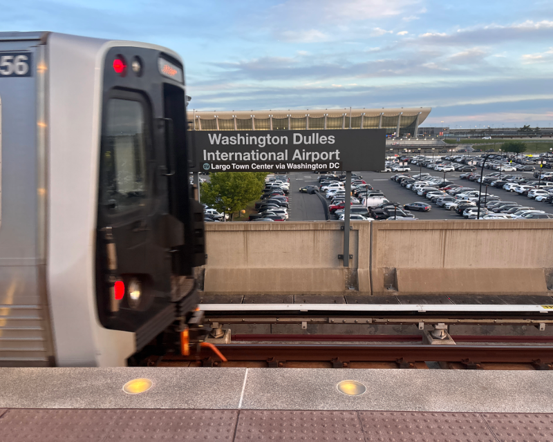 Train pulling away from Washington Dulles International Airport WMATA station with terminal and parking lot in background.