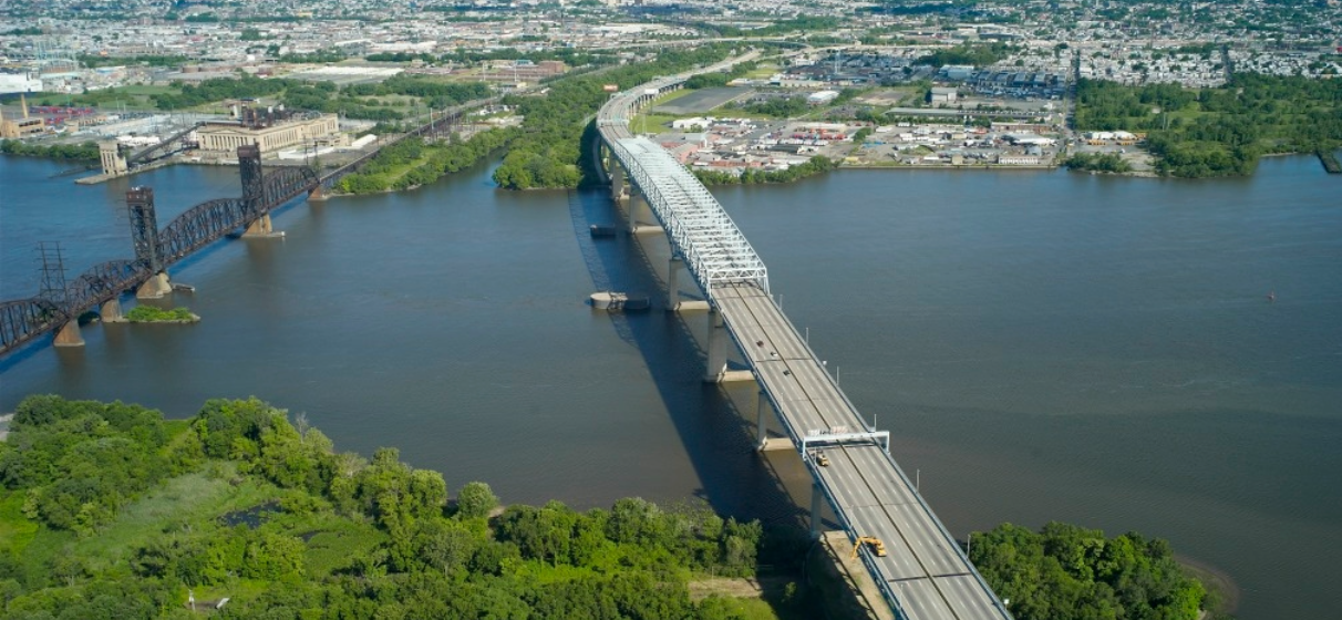 Bird's eye view of highway bridge over water with greenery on one side leading to a city skyline