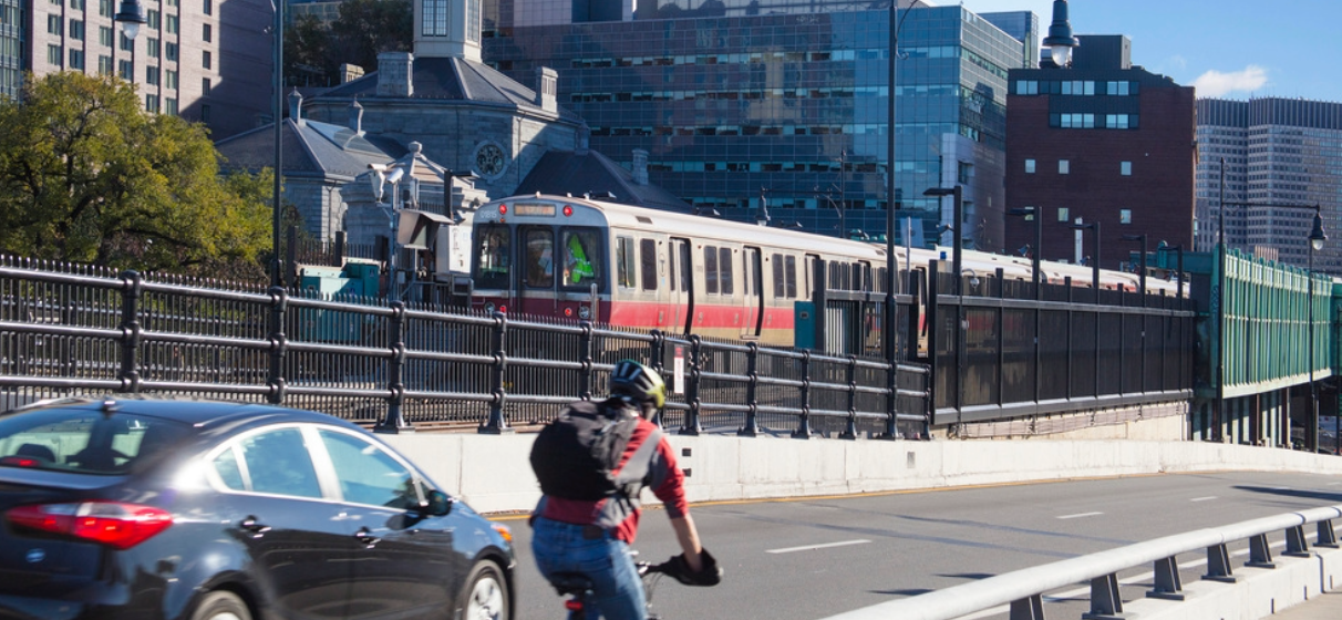 Roadway view of bicyclist, car and train with cityscape background