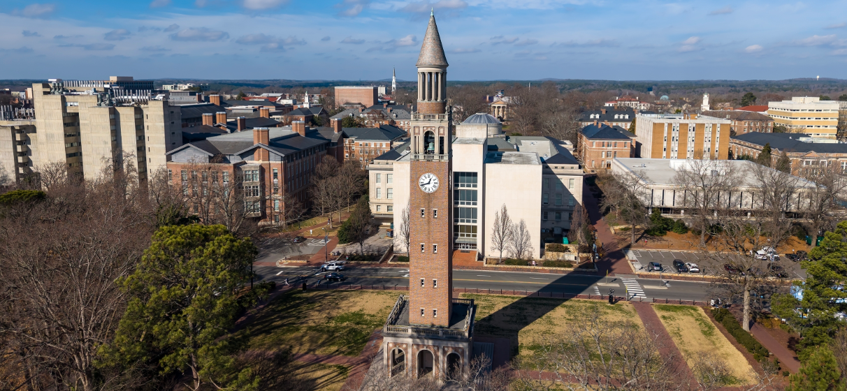 Aerial view of the Morehead-Patterson Bell Tower rises 172 feet above manicured lawns and hedges at UNC Chapel Hill, its Roman numeral clock faces and conical spire marking a timeless campus landmark.