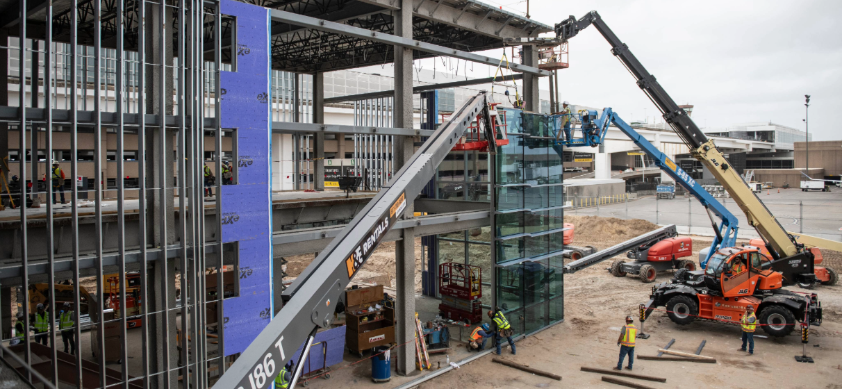 Construction team using cranes and other equipment build the frame of a large airport terminal facility.